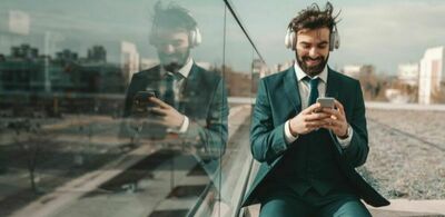 En mann sitter utendørs i dress. Han har på et headset og holder telefonen sin. Foto